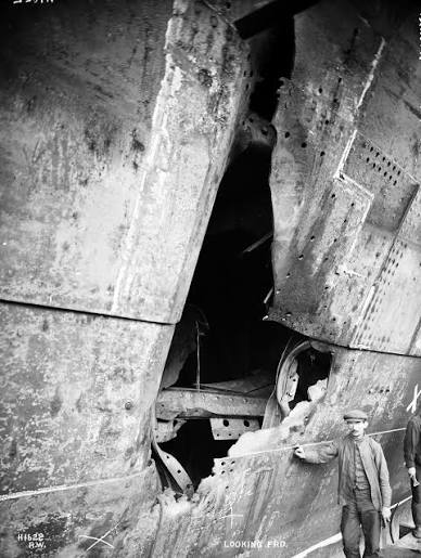 The hull of RMS Olympic torn open above and below the waterline after her 1911 collision with HMS Hawke, a shipyard worker standing at the foot of the damage for scale